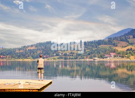 Young man in shorts and vest fishing standing on a floating wooden pier. Stock Photo