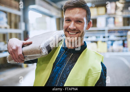 Friendly happy warehouse worker in a high visibility jacket standing in the warehouse with a product bag over his shoulder smili Stock Photo
