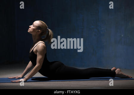 Beautiful sporty fit young woman in black sportswear working out indoors against grunge dark blue wall. Model doing cobra asana Stock Photo