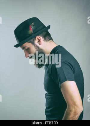 Side view of sad hunched bearded man with hat looking down. Retro toned filtered portrait over gray background with vignette Stock Photo