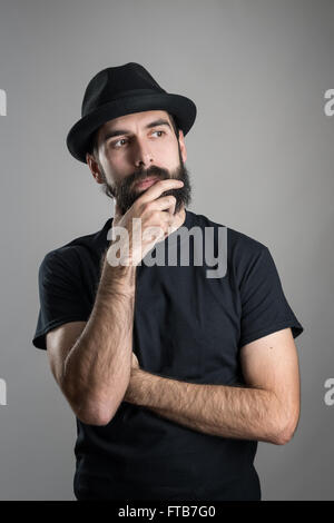 Thinking hipster wearing black t-shirt and hat stroking beard looking away. Headshot portrait over gray studio background Stock Photo