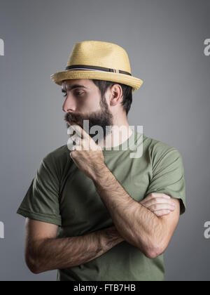 Profile view of young pensive bearded man wearing straw hat touching his beard. Headshot portrait over gray studio background Stock Photo