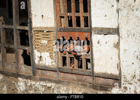 Children poke their heads out of a carved wooden window frame in their dilapidated school building in Nimshong village, Bhutan Stock Photo