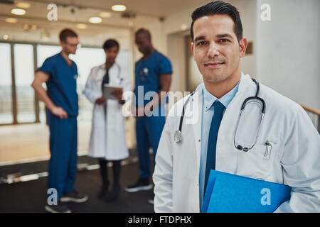 Smiling male doctor wearing white jacket and stethoscope with three diverse colleagues in background Stock Photo