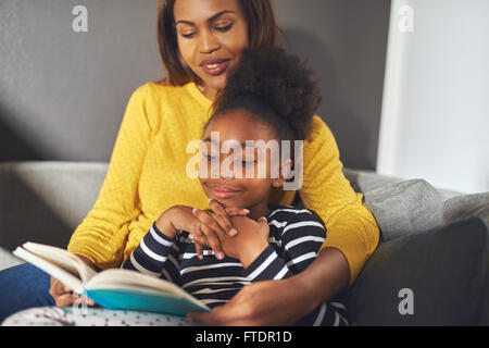 Black mom and daughter reading a book sitting on sofa smiling Stock Photo