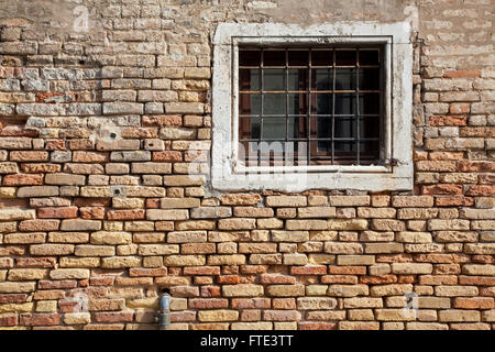 A sunlit barred stone-framed window in a wall of warm coloured crumbling brickwork on Fondamenta Nani, Venice, Italy Stock Photo