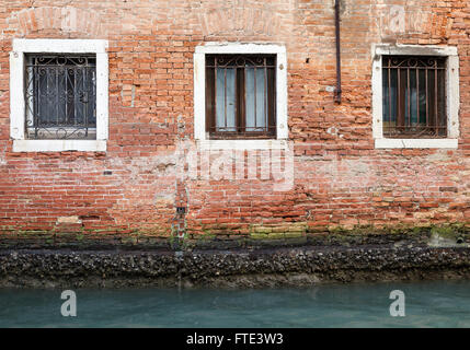Three wrought iron barred stone-framed windows set in crumbling brickwork, green with algae, in the canal side wall of a building in Venice, Italy Stock Photo