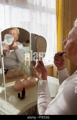 Elderly lady sat at an old fashioned dressing table applying lipstick. Stock Photo