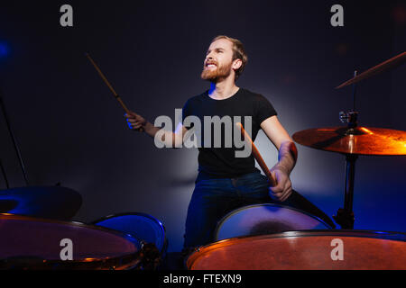Brutal excited young man drummer with beard playing drums over dark background Stock Photo