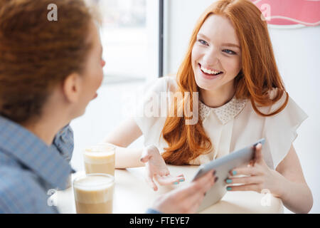 Two cheerful charming young woman smiling and using tablet in cafe Stock Photo