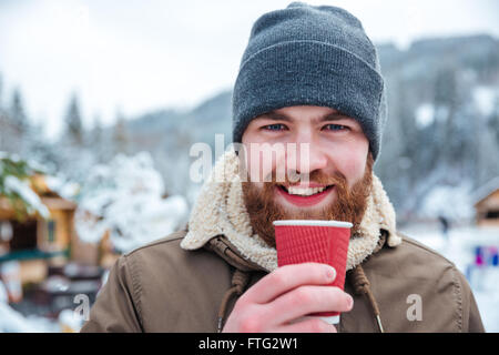 Portrait of cheerful attractive bearded young man drinking hot coffee outdoors in winter Stock Photo