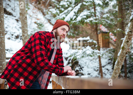 Pensive bearded young man in red hat and checkered shirt standing in winter forest Stock Photo