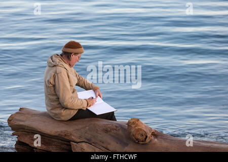Man writing in notebook while sitting on log at edge of Pacific Ocean-Victoria, British Columbia, Canada. Stock Photo