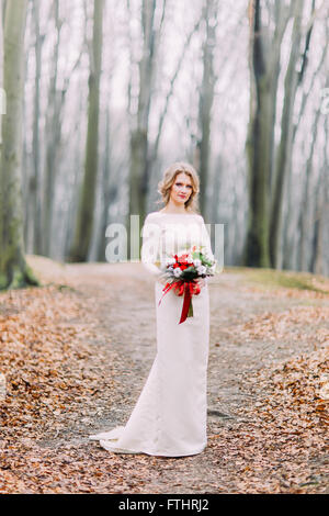 Happy beautiful blonde bride on the autumn forest with bouquet of red flowers Stock Photo