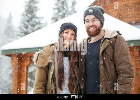 Smiling young couple standing in front of log cabine in winter Stock Photo