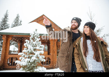 Handsome smiling bearded young man stading with his girlfriend near log cabin and pointing away Stock Photo