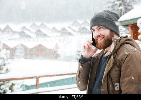 Smiling attractive young man with beard talking on cell phone outdoors in snowy weather Stock Photo