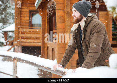 Smiling bearded man in winter wear standing near wooden cottage in winter Stock Photo