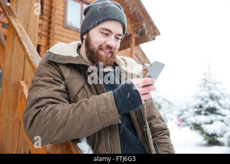 Smiling bearded young man standing and using smartphone outdoors in snowy weather in winter Stock Photo