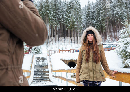 Smiling beautiful young woman standing and posing in winter forest while her boyfriend taking pictures of her Stock Photo