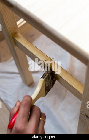 Casual clothed young woman uses a paintbrush to paint with varnish a pine chair or table surface. Stock Photo