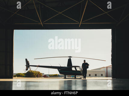 Silhouette of a pilot walking in hangar with a parked helicopter. Pilot arriving at the airport with a helicopter in background. Stock Photo