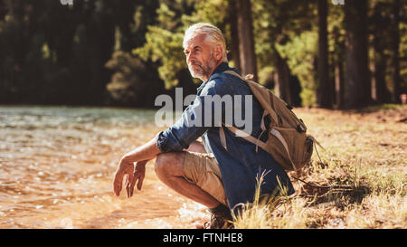 Portrait of a senior man with backpack sitting next to a lake admiring the view. Mature man taking a break from his hike and loo Stock Photo