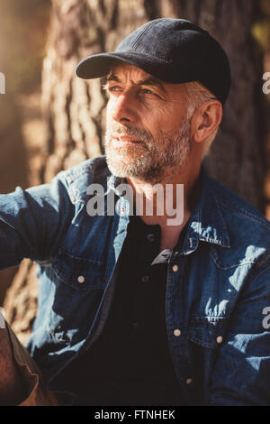 Close up portrait of mature man wearing cap looking away while sitting by a tree. Senior man with beard outdoors on a summer day Stock Photo