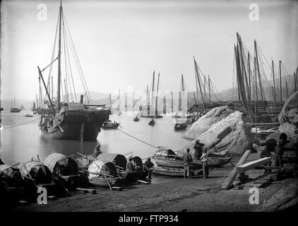 AJAXNETPHOTO. 1890 (APPROX). HONG KONG. A SCENE WITH A LARGE TRADING JUNK MOORED STERN-TO THE SHORE, WITH SMALLER FISHING SAMPANS DRAWN UP ON THE BEACH. TO THE RIGHT ON A ROCK, A CATCH OF FISH DRIES IN THE SUN WHILE A CARPENTER SHIPWRIGHT HAS JUST FINISHED CUTTING A LARGE TIMBER, POSSIBLY FOR NEARBY JUNK BUILDING.   PHOTO:AJAX VINTAGE PICTURE LIBRARY  REF:JUNKS 01 Stock Photo