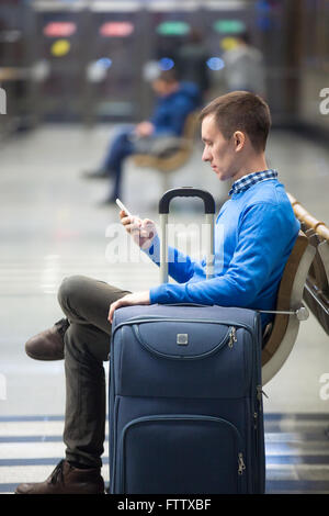 Portrait of young man sitting in airport terminal, holding cellphone. Traveler using public wifi, smartphone app, messaging Stock Photo