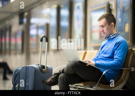 Portrait of young handsome man wearing casual style clothes sitting on the bench in modern airport using laptop. Passenger Stock Photo