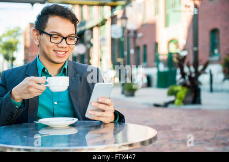 Young asian man in business casual attire sitting and smiling in relaxing outdoor cafe drinking cup of coffee while using mobile Stock Photo