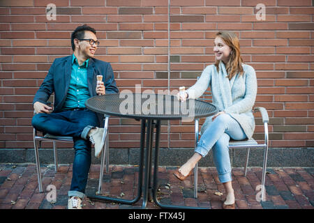 Two smiling young people with ice cream sitting at outdoor cafe table near brick wall having conversation Stock Photo