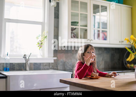 Girl sitting in kitchen eating chocolate dessert and licking fingers Stock Photo