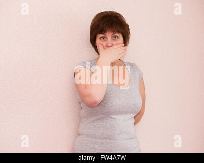 Portrait attractive mature woman with shocked, anxious facial expression, covering mouth with hand on plain background Stock Photo