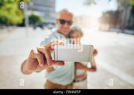 Happy senior couple embracing and taking a self portrait on mobile phone outdoors. Tourist taking selfie, focus on mobile phone. Stock Photo