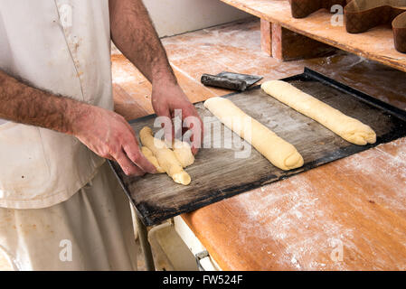 Baker working with uncooked dough forming it into long rolls and shapes, close up of his hands and a long wooden table Stock Photo