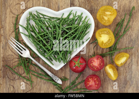 Diet health food of samphire and tomato vegetables on a heart shaped china plate with silver fork and loose over old wood. Stock Photo