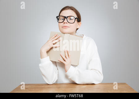 Happy pretty teenage girl in glasses sitting and holding book over gray Stock Photo