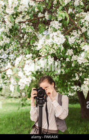 Woman shoots at the camera on a tripod, monopod against a background of trees, park, apple blossom Stock Photo