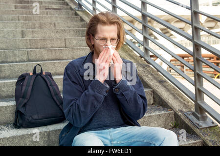 young man with long red hair and glasses sitting outside on stairs and blowing his nose Stock Photo
