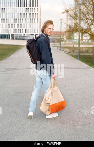 young man with long red hair, beard and glasses walking in the street with a backpack Stock Photo
