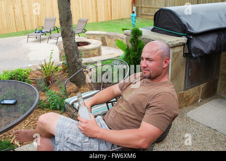 Cute bald muticultural man sitting on a patio in the back yard by bbq grill near his cell phone drinking a beer. Stock Photo
