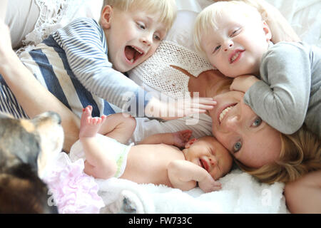 A happy young mother is laying on the bed with her three young children, two wild boys and a newborn baby girl, as their pet dog Stock Photo