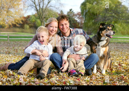A happy family of four people, including mother, father, young child, and toddler brother are sitting outside in fallen leaves Stock Photo