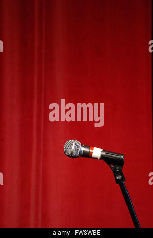 A microphone stands alone in a spotlight on stage in front of a red curtain. Stock Photo