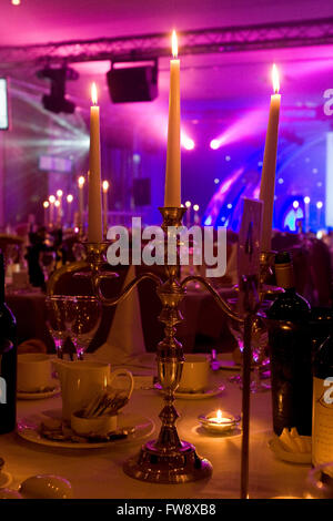 A dining hall in a large hotel that has been fitted out for a corporate evening event, atmospheric lighting compliments the dining area where tables are set out with candles and also a stage is lit up in bright colours at the far end of the room. Stock Photo
