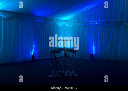 An empty bar area in a tent set up at a function for business presentations and annual general meetings. Coloured with rich blue light the room is empty but for a small table with four bar stools arranged aropund it. Stock Photo