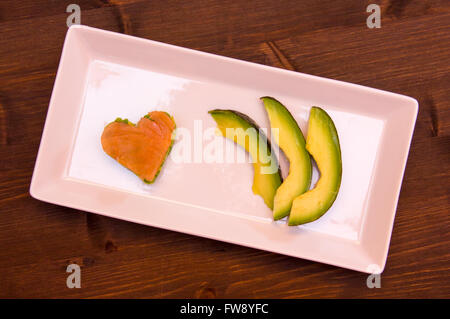 Slices of avocado and avocado and smoked salmon in a heart shape on the tray on wood seen from above Stock Photo