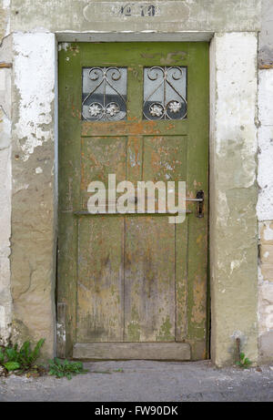 Old, green, weathered wooden door with two small barred glass panes in the dilapidated facade of an abandoned house. Stock Photo